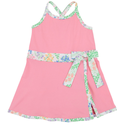 Girls’ Prepletic™ Teggy Tennis Dress by The Beaufort Bonnet Company—a pink sleeveless dress with a floral trim and matching waist sash, featuring a crossover back and floral edging at the hem and neckline for a playful yet polished activewear-inspired look.