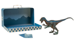 The Coco Village Velociraptor Dinosaur in a Collectible Box is pictured beside a blue checker-patterned suitcase featuring a mountain-themed interior and two cards, creating an imaginative prehistoric scene on a white background.