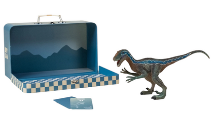 The Coco Village Velociraptor Dinosaur in a Collectible Box is pictured beside a blue checker-patterned suitcase featuring a mountain-themed interior and two cards, creating an imaginative prehistoric scene on a white background.