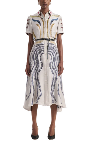 A woman wears the Kiera Dress by Altuzarra, a white A-line button-down with short sleeves and colorful abstract patterns, against a white background.