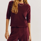 A person wearing a slim-fitting maroon Corfu Velour Top with three-quarter sleeves and matching tapered pants.