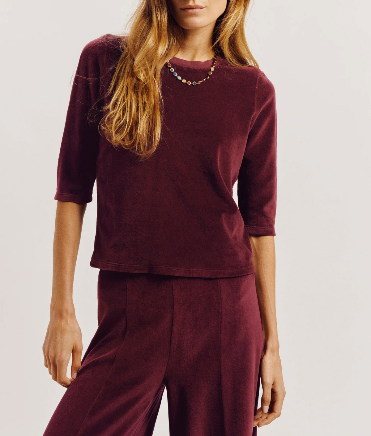 A person wearing a slim-fitting maroon Corfu Velour Top with three-quarter sleeves and matching tapered pants.