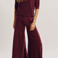 A person wearing a dark red, slim-fitting Corfu Velour Top and matching wide-leg trousers that emphasize a tapered body silhouette.
