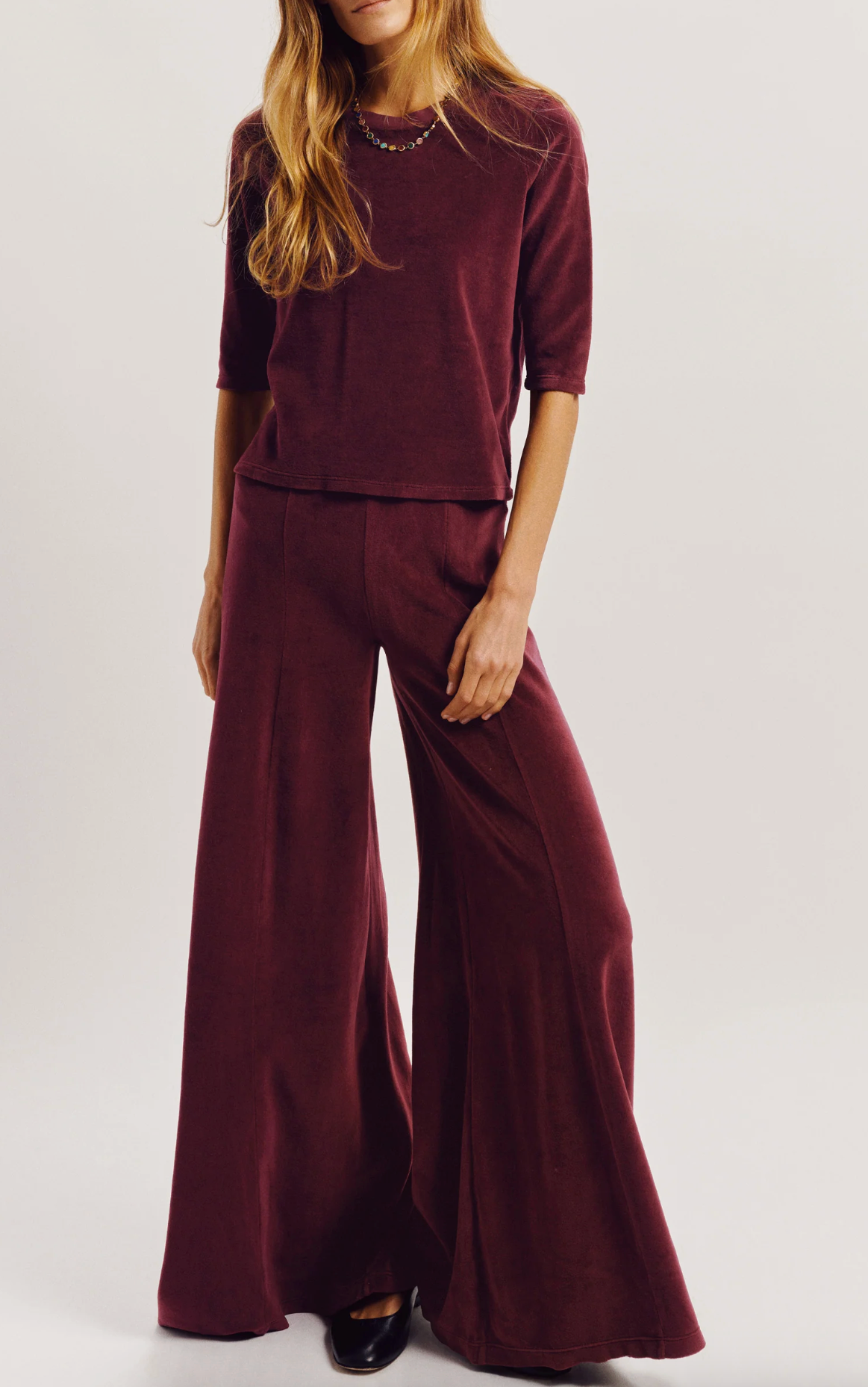 A person wearing a dark red, slim-fitting Corfu Velour Top and matching wide-leg trousers that emphasize a tapered body silhouette.
