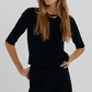 A woman wearing a black, slim-fitting, three-quarter sleeve Suzie Kondi Corfu Velour Top with a gold chain collar.
