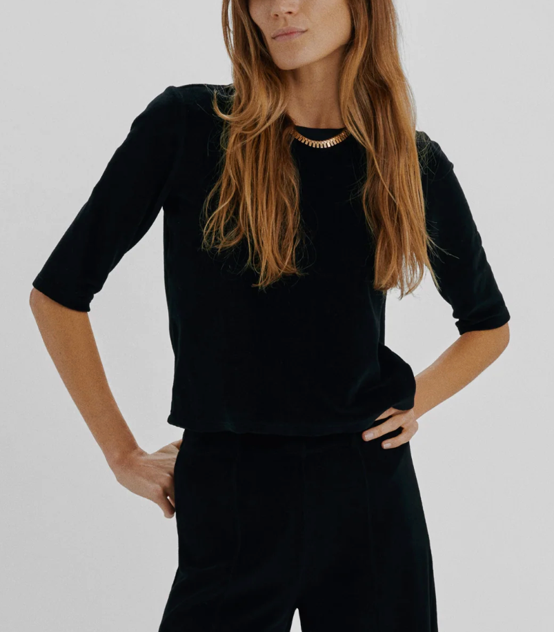 A woman wearing a black, slim-fitting, three-quarter sleeve Suzie Kondi Corfu Velour Top with a gold chain collar.