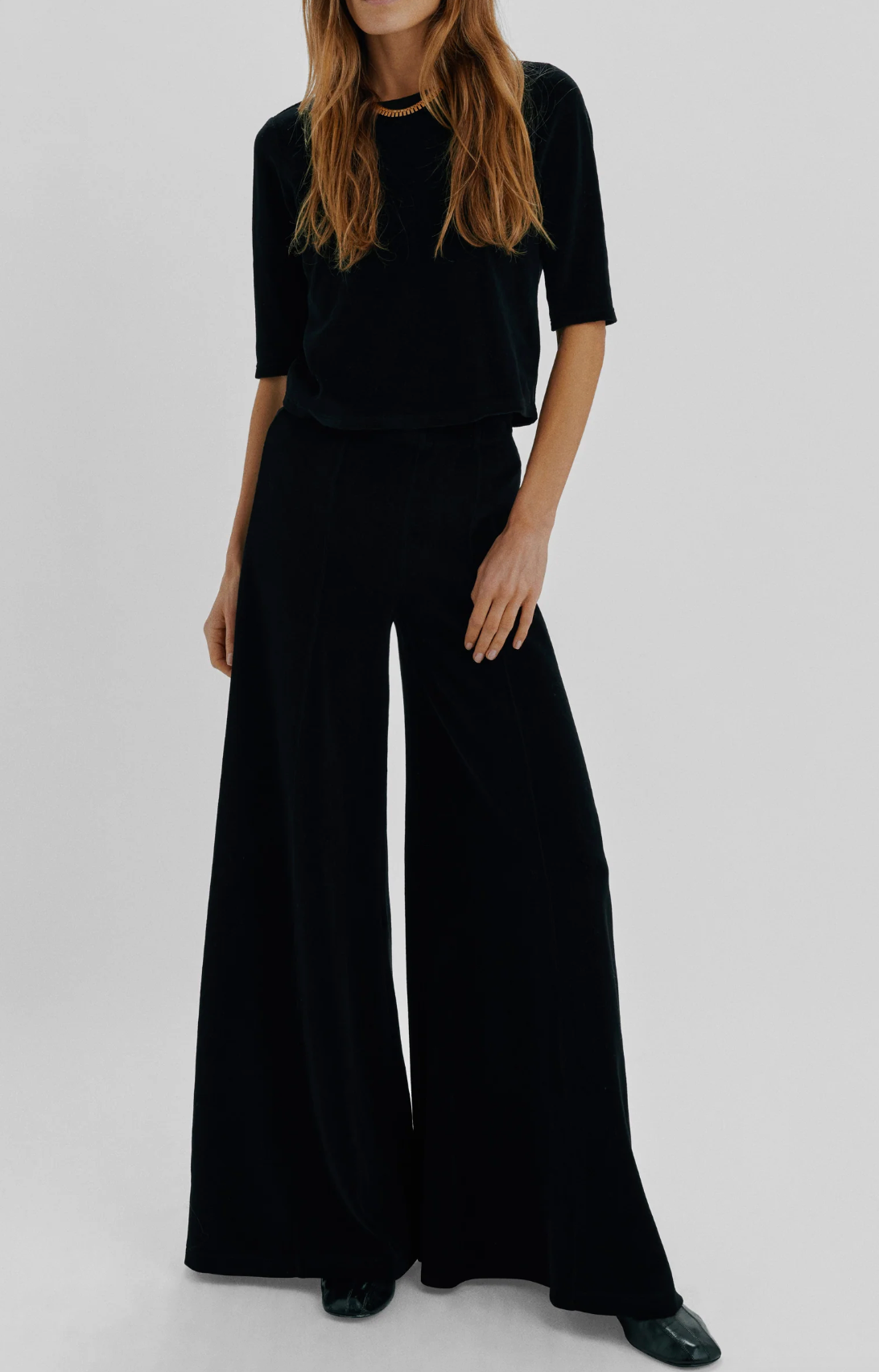 A person wearing a short-sleeved black top and black Suzie Kondi Megalo Velour Pants with a front seam.
