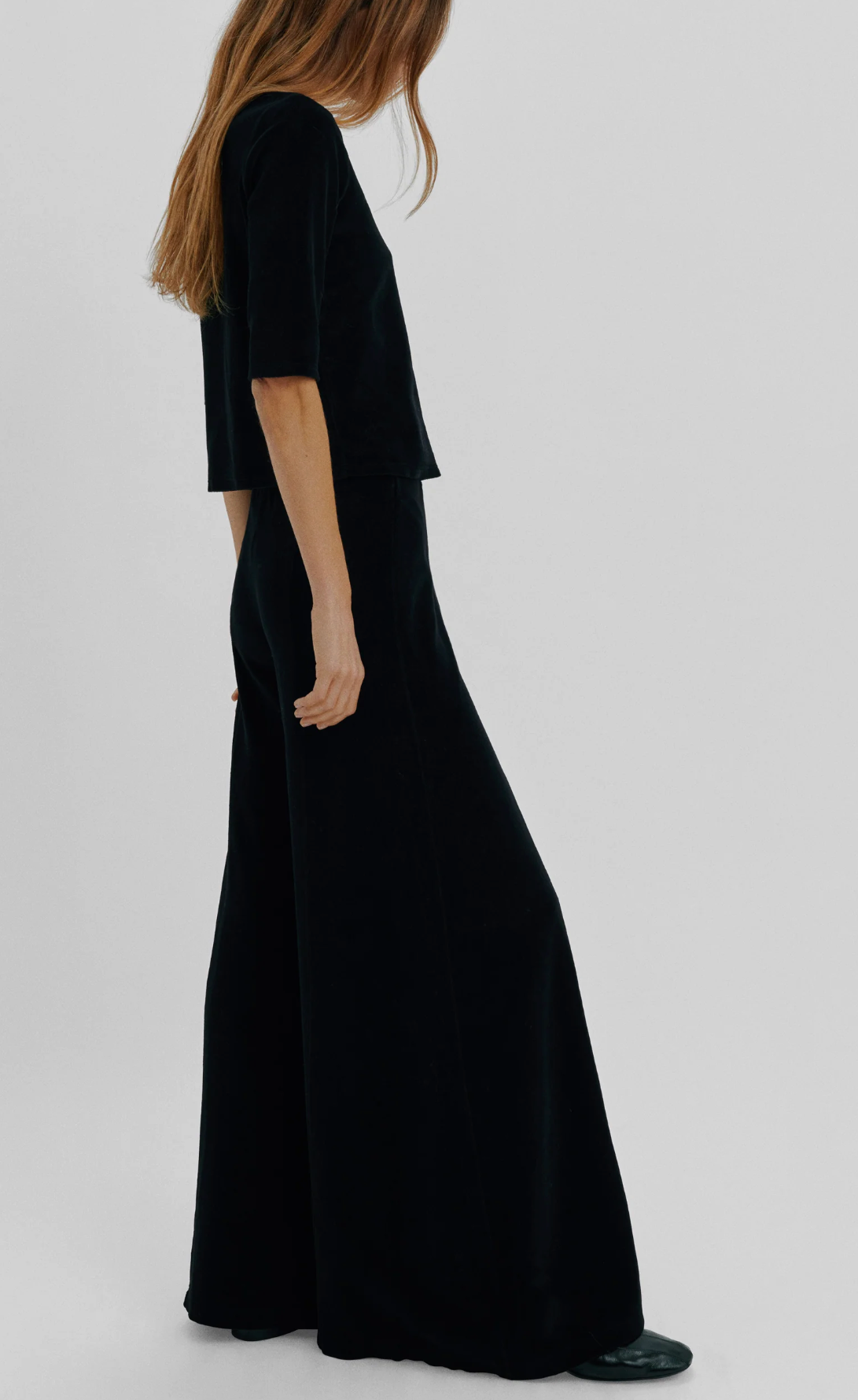 A person wearing a black short-sleeved top and Suzie Kondi Megalo Velour Pants, featuring a wide-leg flared silhouette and front seam.