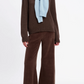 A model wearing a brown crewneck sweater, Suzie Kondi Zephyra Velour Pants in a relaxed wide-leg fit, and a light blue sweater draped over their shoulders.