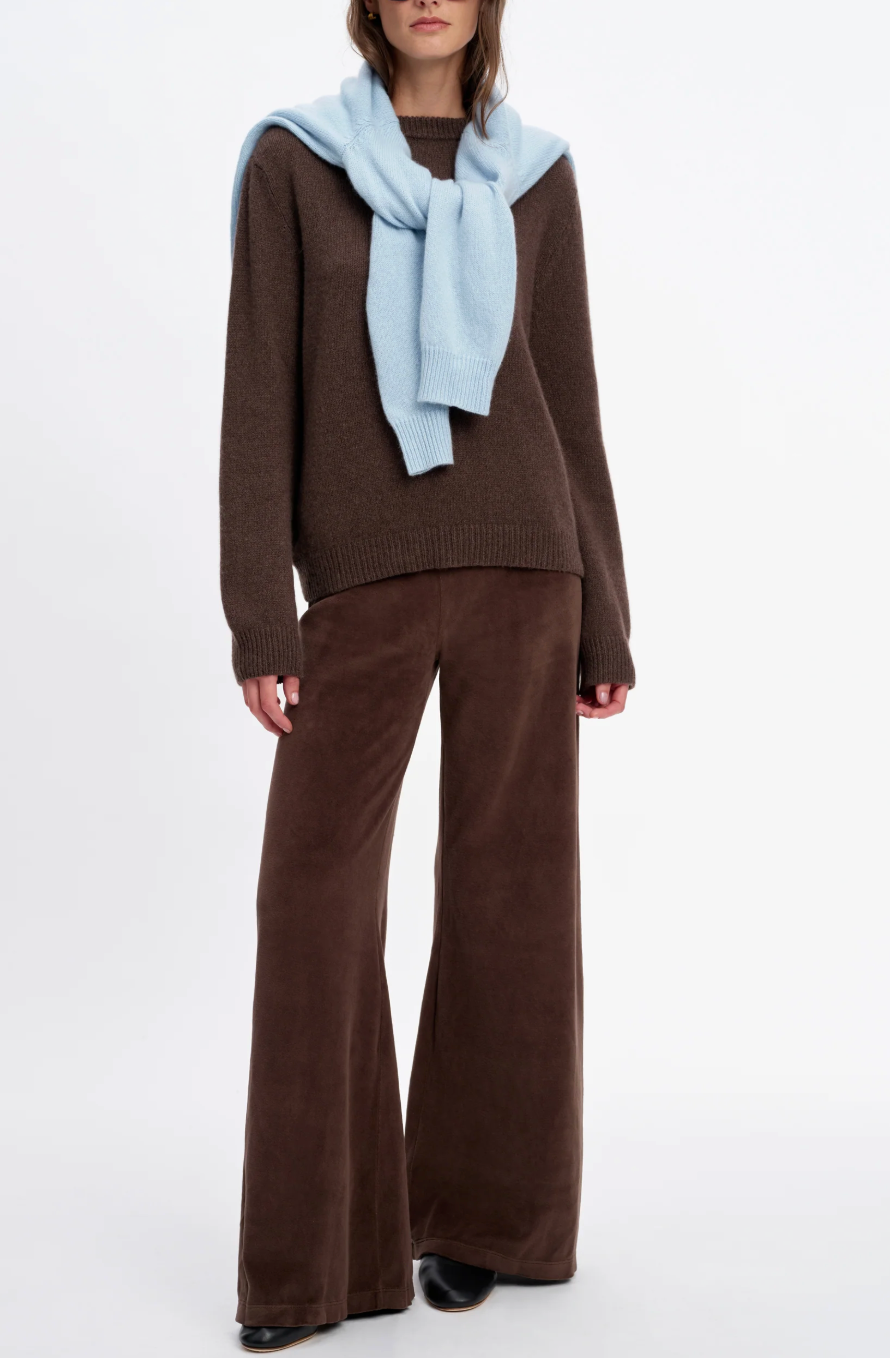 A model wearing a brown crewneck sweater, Suzie Kondi Zephyra Velour Pants in a relaxed wide-leg fit, and a light blue sweater draped over their shoulders.
