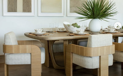 neutral dining room with light wood table, beige and light wood chairs, and white table settings with palm front centerpiece