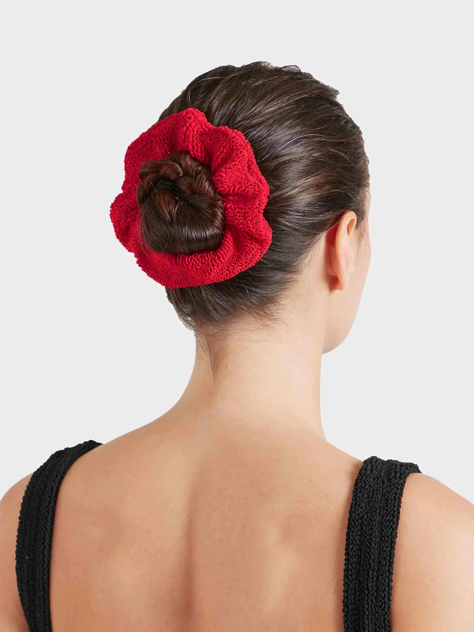 A woman with dark hair in a bun, secured by the Hunza G Scrunchie, wears a black sleeveless textured top and is shown from the back against a plain background.