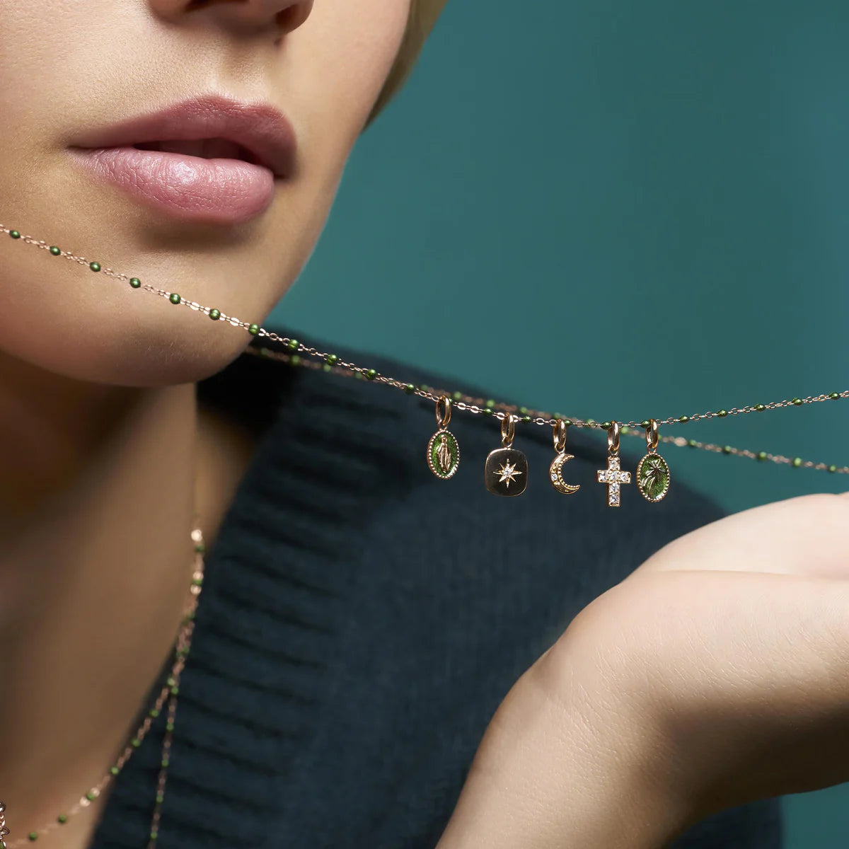 A person holds the Gigi Clozeau Cross Diamond Pendant, showcasing its timeless design with various small pendants like a star, crescent moon, and cross against a blue-green background.