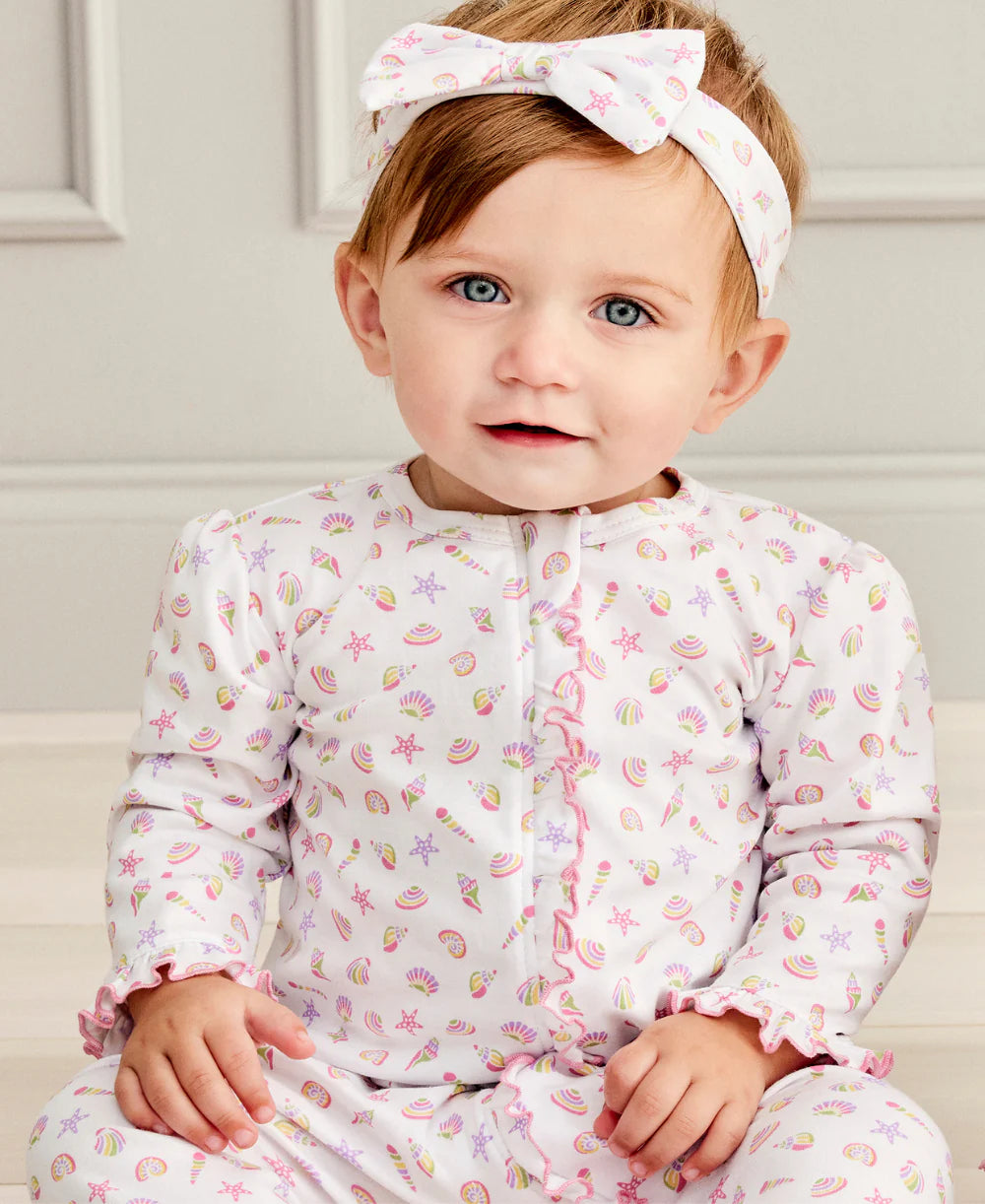 A baby wears the Kissy Kissy Baby Girls' Sea Shell Magic Zip Footie in soft Pima cotton with pastel seashell and starfish print, paired with a matching white headband, sitting indoors against a light background.
