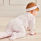 A baby girl in the Kissy Kissy Baby Girls' Sea Shell Magic Zip Footie crawls on a light-colored floor with beige paneled walls, adding a touch of sea shell magic to the cozy room.