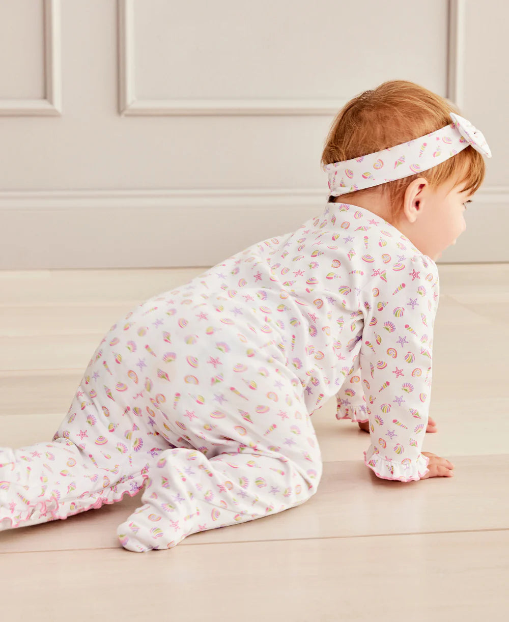A baby girl in the Kissy Kissy Baby Girls' Sea Shell Magic Zip Footie crawls on a light-colored floor with beige paneled walls, adding a touch of sea shell magic to the cozy room.