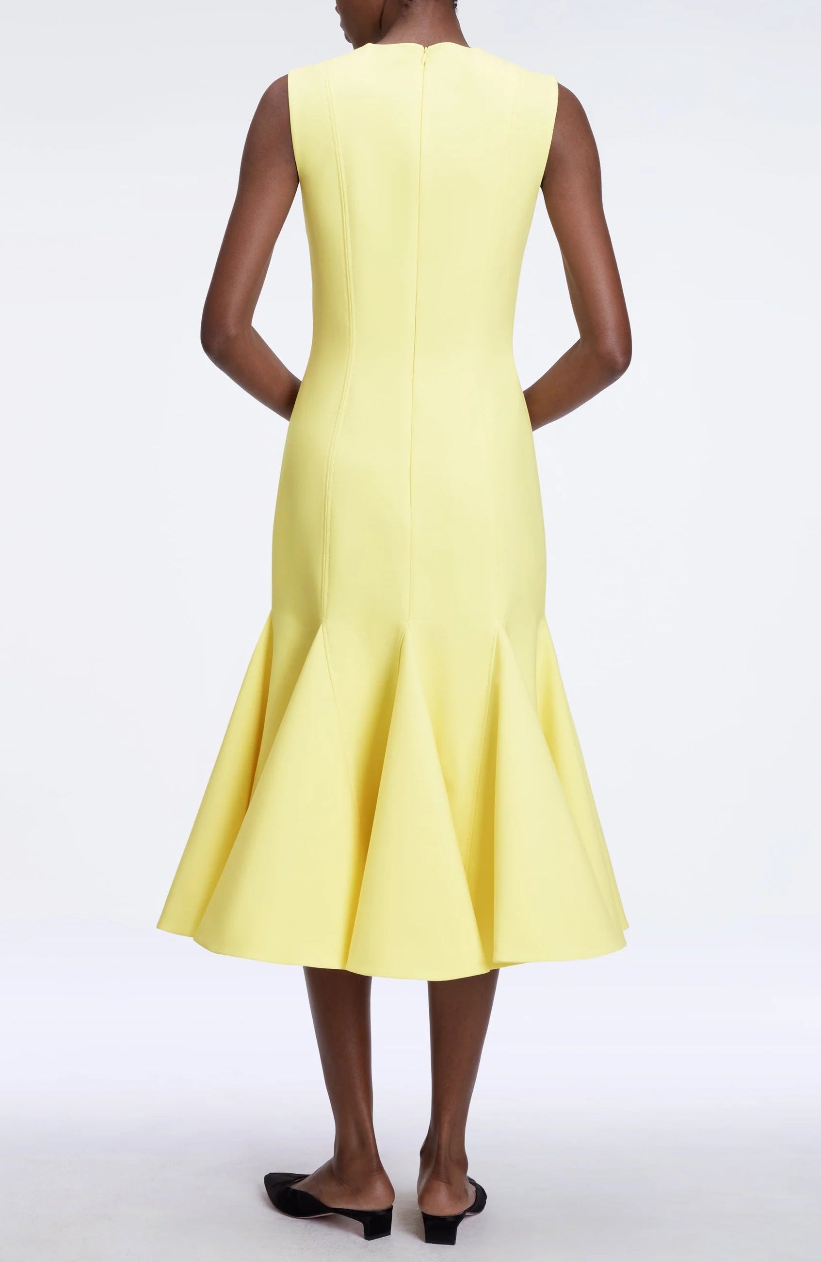 A person stands with hands behind their back, facing away from the camera, wearing the Seema Dress by Emilia Wickstead—a sleeveless, fitted yellow summer wool dress with a flared hem.