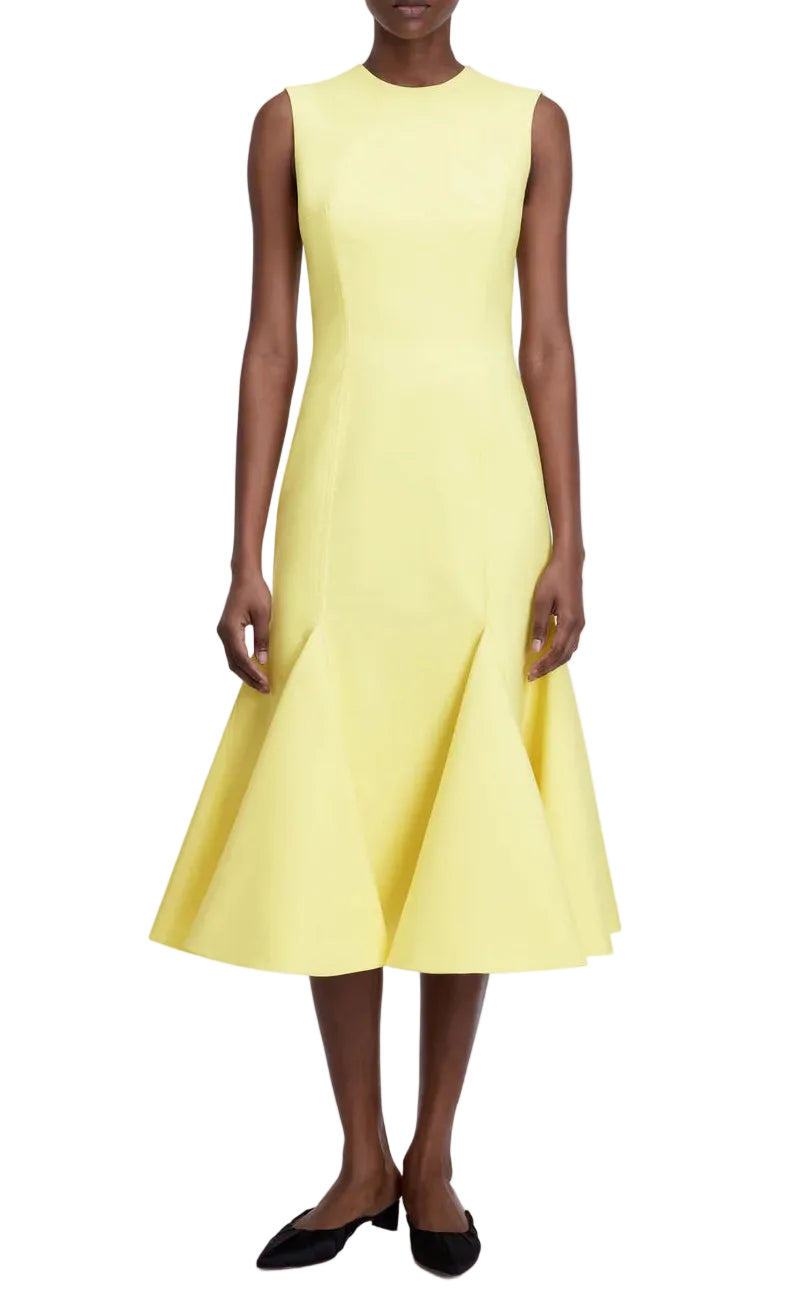 A woman wearing the Seema Dress by Emilia Wickstead, a sleeveless yellow wool dress with a fitted bodice and flared hem, paired with black flats, stands against a white background.