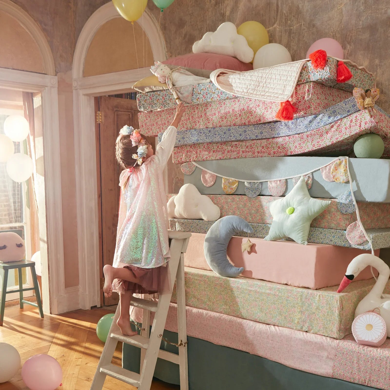 A child in a playful outfit, complete with the Meri Meri Iridescent Sequin Cape & Wand, climbs a ladder to reach a stuffed cat toy on top of a tall stack of colorful mattresses, surrounded by balloons and plush decorations in a whimsical room.