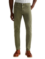 A person wearing AG Jeans Tellis Modern Slim pants in green Italian sateen fabric and brown boots stands with their legs shoulder-width apart against a plain background.