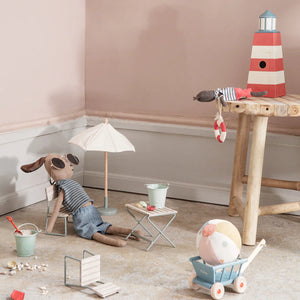 Maileg stuffed animals relax with a Miniature Sunshade, toy beach furniture, a garden set, a wagon and beach ball, plus a wooden lighthouse in a pastel-walled corner room.