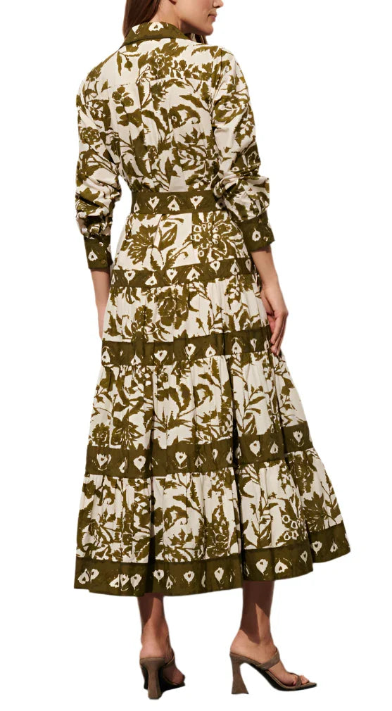 A woman is depicted from behind wearing a Figue Shelby Dress, which boasts a long-sleeved, floral-patterned design in shades of white and green. The dress highlights a voluminous tiered skirt and she pairs it elegantly with open-toe heeled sandals.