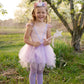A young girl wearing the Great Pretenders Shimmer Unicorn Dress & Headband, complete with a pink fairy costume featuring a tulle skirt and holding a star wand, stands on grass near blossoming trees.