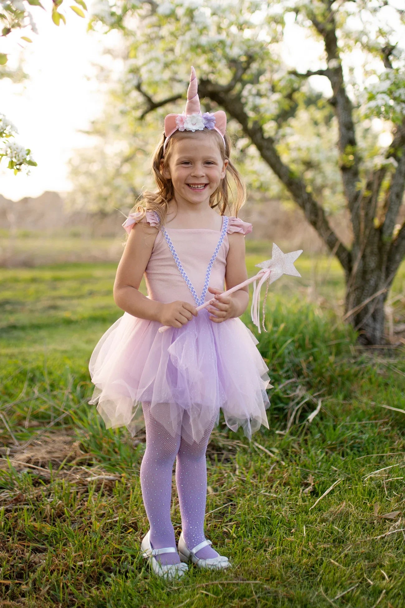 A young girl wearing the Great Pretenders Shimmer Unicorn Dress & Headband, complete with a pink fairy costume featuring a tulle skirt and holding a star wand, stands on grass near blossoming trees.