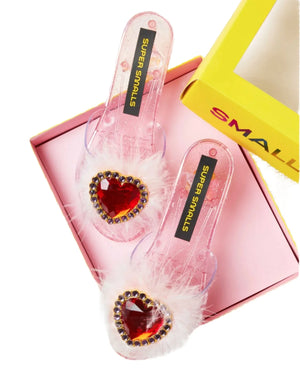 A pair of Super Smalls Daydream Queen Play Shoes, designed with pink transparent material adorned with heart-shaped gems, white feather accents, and a glitter design, showcased in an opened yellow box. Perfect for a dress-up debut as the ultimate Daydream Queen!