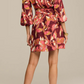 A woman stands facing away, wearing the Figue Izzie Dress—a pink, yellow, and white abstract-patterned band collar, long sleeve A-line dress—paired with wedge heels.
