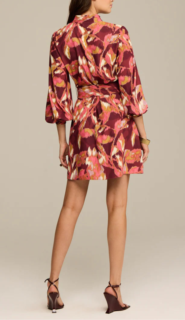 A woman stands facing away, wearing the Figue Izzie Dress—a pink, yellow, and white abstract-patterned band collar, long sleeve A-line dress—paired with wedge heels.