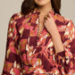 A woman in the Figue Izzie Dress, featuring a band collar, matte crepe in maroon, pink, and white, and a buttoned neckline, stands with one hand to her neck. Wide sleeves and an A-line silhouette complete the elegant look.