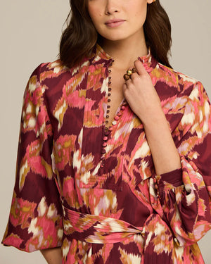 A woman in the Figue Izzie Dress, featuring a band collar, matte crepe in maroon, pink, and white, and a buttoned neckline, stands with one hand to her neck. Wide sleeves and an A-line silhouette complete the elegant look.
