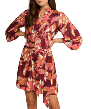 A woman wears the Figue Izzie Dress, a maroon matte crepe piece with pink, orange, and white abstract floral prints, long sleeves, a belted waist, and an A-line knee-length silhouette.