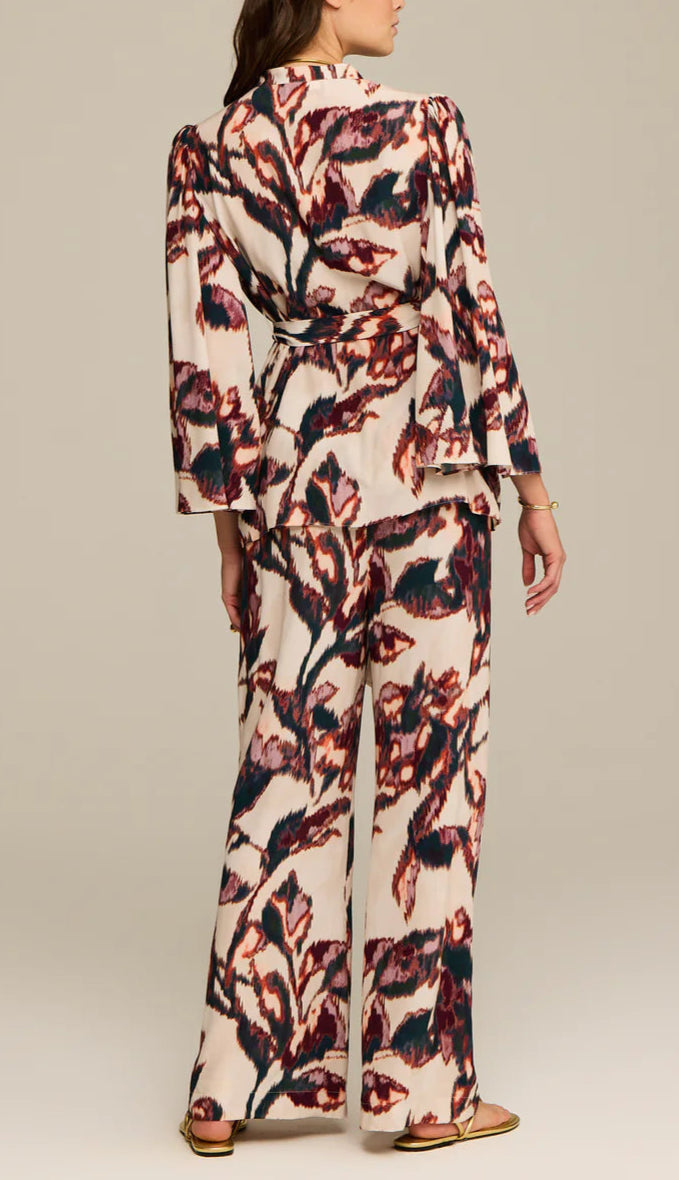 A woman faces away, wearing the Figue Avery Top—a loose, long-sleeve viscose crepe shirt with an abstract multicolor floral print—paired with flat sandals.