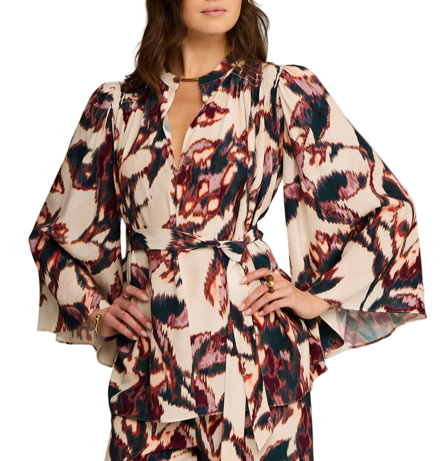 A woman wears the Avery Top by Figue—a long-sleeve, patterned viscose crepe blouse featuring a deep V-neck, tie waist, and butterfly sleeves in red, black, and cream.