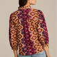 A person is shown from the back wearing the Figue Danilia Top, a long-sleeve, band collar blouse featuring a geometric print in purple, orange, and yellow.