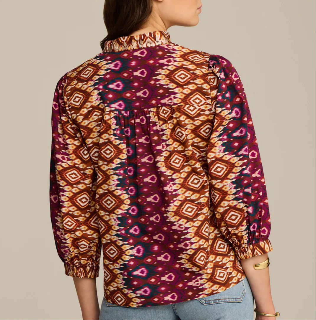 A person is shown from the back wearing the Figue Danilia Top, a long-sleeve, band collar blouse featuring a geometric print in purple, orange, and yellow.