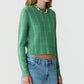 A person wearing the Guest in Residence Shrunken Checker Crew—a green, long-sleeve women’s cashmere sweater with a checkered pattern—paired with blue jeans and a beaded necklace, standing against a plain light background.