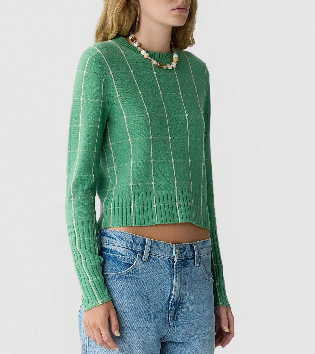 A person wearing the Guest in Residence Shrunken Checker Crew—a green, long-sleeve women’s cashmere sweater with a checkered pattern—paired with blue jeans and a beaded necklace, standing against a plain light background.