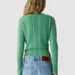 A woman is facing away, wearing the Guest in Residence Shrunken Checker Crew—a green, long-sleeve women’s cashmere sweater with a grid pattern—paired with light blue high-waisted jeans featuring a brown leather patch on the waistband.