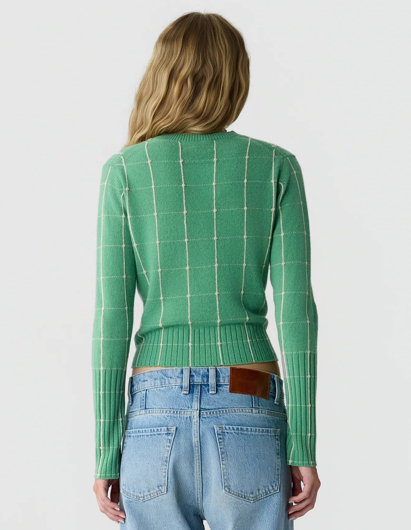 A woman is facing away, wearing the Guest in Residence Shrunken Checker Crew—a green, long-sleeve women’s cashmere sweater with a grid pattern—paired with light blue high-waisted jeans featuring a brown leather patch on the waistband.