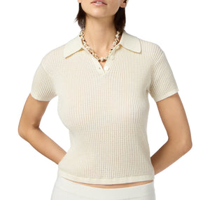 A woman wears the Guest in Residence Shrunken Cashmere Polo in off-white, featuring a lightweight waffle knit, short sleeves, and a collar. She pairs it with a beaded necklace and stands with her arms behind her back.