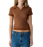A woman stands against a white background wearing the Guest in Residence Shrunken Waffle Polo—a brown, short-sleeve, collared crochet knit top with a subtle waffle texture reminiscent of a women’s cashmere polo—paired with blue jeans and a pearl necklace.