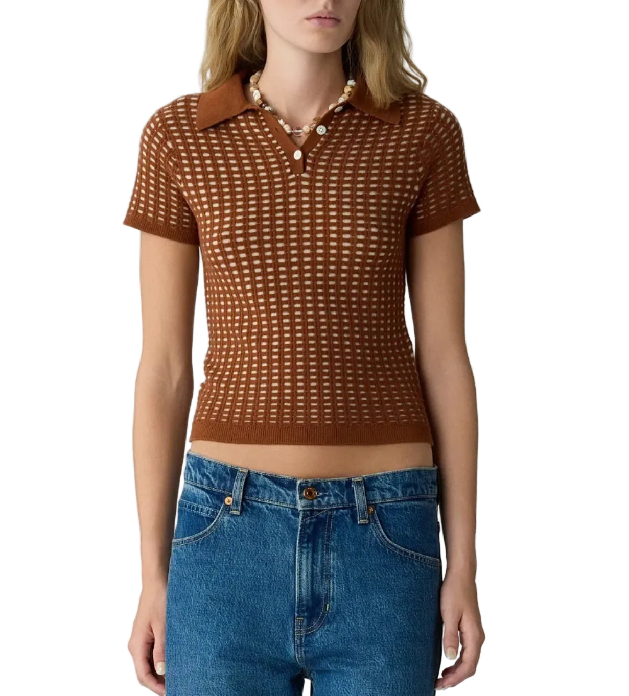 A woman stands against a white background wearing the Guest in Residence Shrunken Waffle Polo—a brown, short-sleeve, collared crochet knit top with a subtle waffle texture reminiscent of a women’s cashmere polo—paired with blue jeans and a pearl necklace.