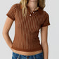 A woman wearing the Guest in Residence Shrunken Waffle Polo—a brown, short-sleeve women’s cashmere shirt with a grid pattern—paired with blue jeans, standing against a plain light background.