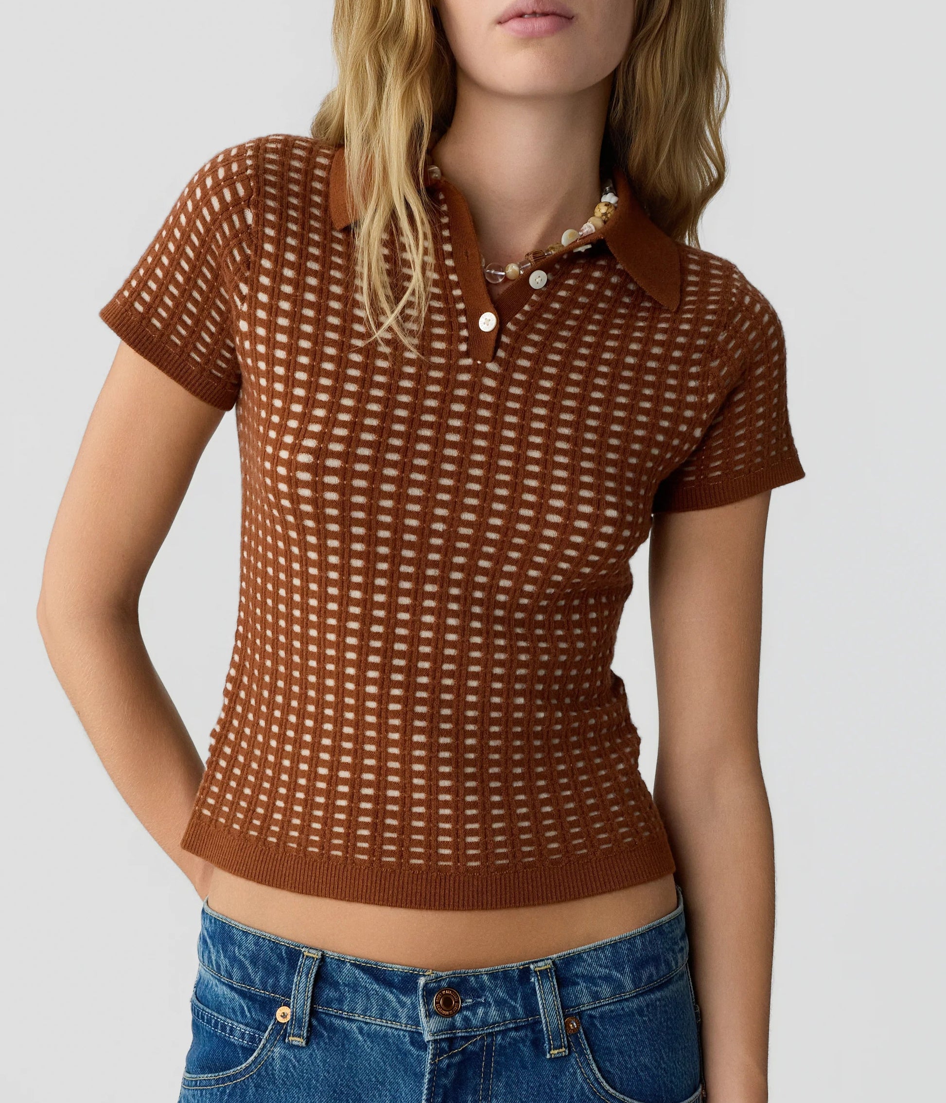 A woman wearing the Guest in Residence Shrunken Waffle Polo—a brown, short-sleeve women’s cashmere shirt with a grid pattern—paired with blue jeans, standing against a plain light background.