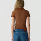 A person with long, wavy blonde hair is shown from the back against a plain light background, wearing Guest in Residence’s Shrunken Waffle Polo—a short-sleeve brown women’s cashmere shirt with a subtle waffle texture—paired with blue jeans.