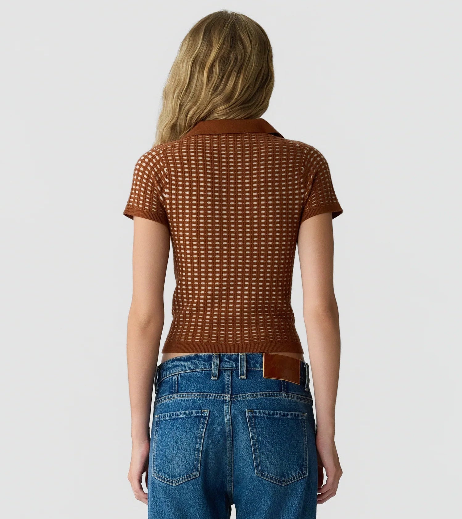 A person with long, wavy blonde hair is shown from the back against a plain light background, wearing Guest in Residence’s Shrunken Waffle Polo—a short-sleeve brown women’s cashmere shirt with a subtle waffle texture—paired with blue jeans.