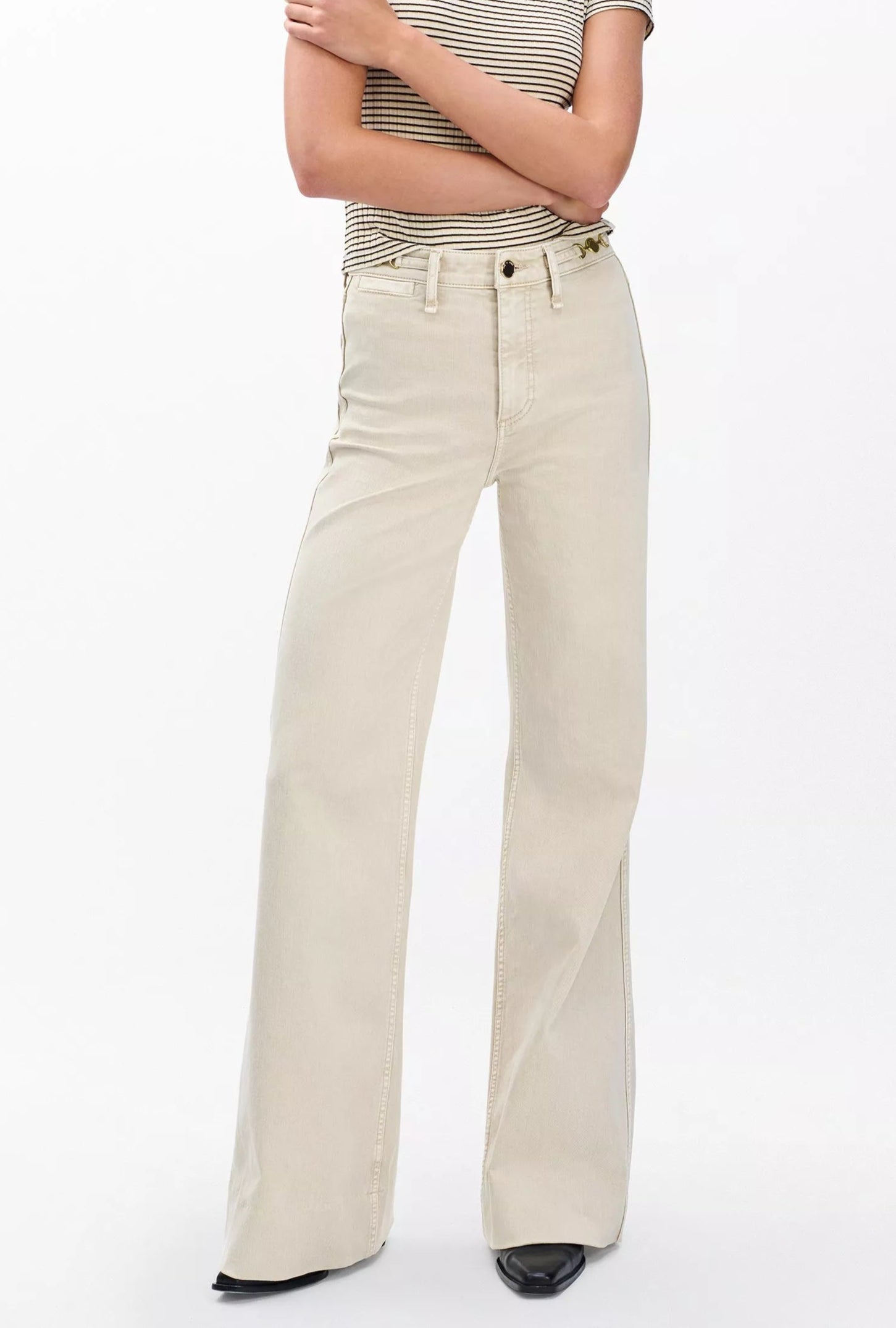 A person wears Rag & Bone's Skylar High Rise Trouser Jean in beige vintage stretch denim, paired with a short-sleeve striped shirt and black pointed shoes, standing against a plain white background.